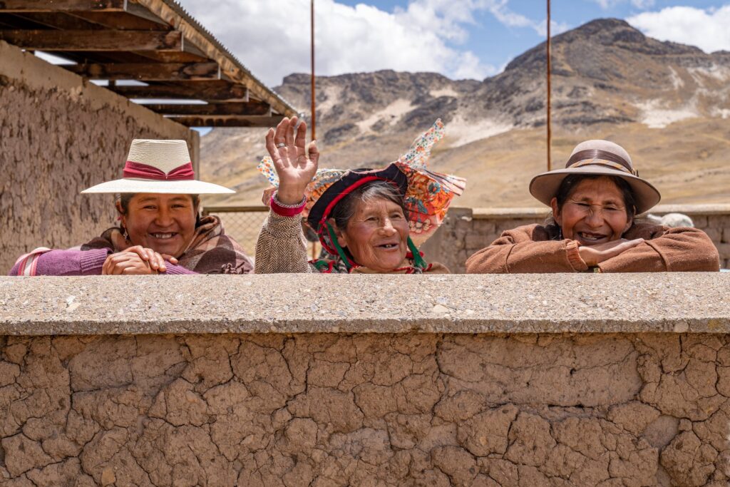 Three Quechua women in traditional dress waving from a wall in the high Andes, Picotani, Peru